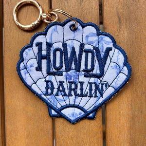 May include: A seashell-shaped keychain featuring a light blue and white floral fabric base with dark blue embroidered edges and the text HOWDY DARLIN stitched in dark blue thread. The keychain includes a gold-toned metal ring attachment.