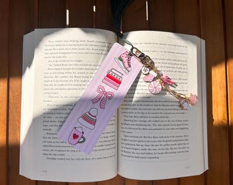 May include: A pink bookmark with winter-themed illustrations, including ice skates, books, a bow, a hat, and a mug. A gold chain with charms, including a letter "W", a pink candy, and a star, hangs from the bookmark.