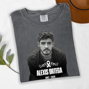 May include: A gray Comfort Colors t-shirt featuring a black and white portrait of a man. Below the portrait is the name "ALEXIS ORTEGA" with the years "1987-2026" and a ribbon graphic.