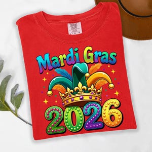 May include: A red t-shirt with a colorful Mardi Gras design. The design includes the words "Mardi Gras" in rainbow colors, a jester hat, a crown, and the year "2026". The shirt is a Comfort Colors brand.