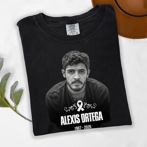 May include: Black t-shirt featuring a black and white portrait of a man with the name "ALEXIS ORTEGA" and the years "1987-2026" printed below. The shirt has a crew neck and the Comfort Colors brand tag.