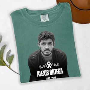 May include: A green Comfort Colors t-shirt featuring a black and white portrait of a man. The shirt includes the text "ALEXIS ORTEGA" with the years "1987-2026" and a white ribbon graphic.