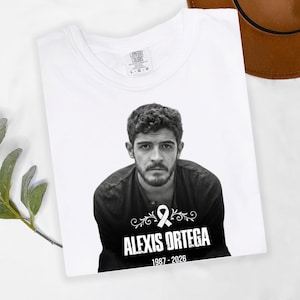 May include: White t-shirt featuring a black and white portrait of a man. The shirt includes the name "ALEXIS ORTEGA" and the years "1987-2026" below the portrait. The shirt is folded and has a tag that says "COMFORT COLORS".