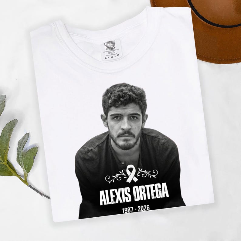 May include: White t-shirt featuring a black and white portrait of a man. The shirt includes the name "ALEXIS ORTEGA" and the years "1987-2026" below the portrait. The shirt is folded and has a tag that says "COMFORT COLORS".