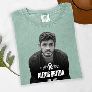 May include: A light green Comfort Colors t-shirt featuring a black and white portrait of a man. The text "ALEXIS ORTEGA 1987-2026" is printed below the portrait, along with a white ribbon graphic.