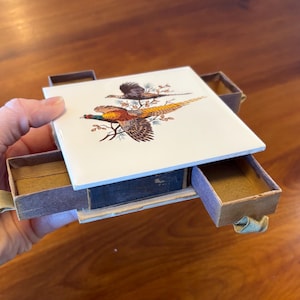 May include: A small, open wooden box with two pull-out drawers. The top of the box is a white tile with a colorful illustration of two pheasants. The drawers are empty and lined with a light brown material.