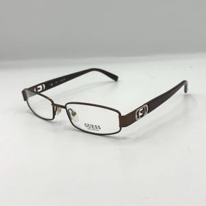 May include: Brown rectangular eyeglasses with the brand name "GUESS" printed on the lens. The frames are a dark brown color with a silver accent on the temples. The glasses have a classic, understated design.