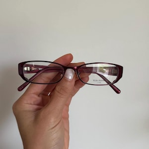 May include: Dark pink Vera Wang Luxe eyeglasses. The rectangular frames have a subtle cat-eye shape and clear lenses. The brand name is visible on the right lens.