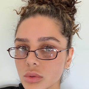 May include: Rectangular eyeglasses with a dark brown frame. The lenses are clear, and the glasses are worn by a person. The frame has a thin, delicate design. The overall style is classic and understated.