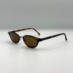 May include: Oval-shaped sunglasses featuring a brown tortoiseshell pattern frame and dark brown tinted lenses. The glasses have thin, matching tortoiseshell temples and a metallic bridge piece connecting the two circular rims.