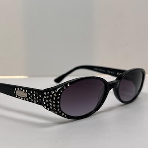 May include: Black oval sunglasses featuring dark tinted lenses and a frame embellished with clear rhinestones along the outer edges. A small silver-toned metal plaque on the temple displays the text Fossil Signature in black lettering.