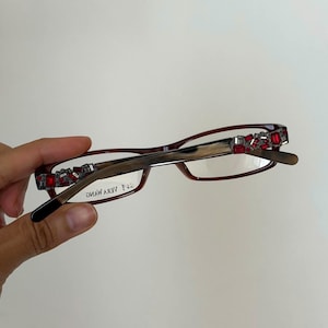 Peut inclure: Lunettes de couleur bordeaux avec des ornements de bijoux rouges décoratifs sur les coins supérieurs. Les montures sont de forme rectangulaire et portent le nom de la marque "VERA WANG" sur le verre.