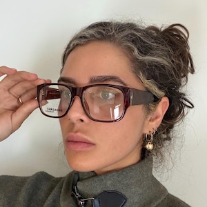 May include: Rectangular eyeglasses with a dark burgundy frame and clear lenses. The temples are a darker shade of brown. The text "BROADWAY" is visible on the lens. The glasses are worn by a person.