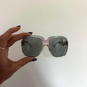 Könnte beinhalten: Übergroße Sonnenbrille mit einem hellblauen und rosa Farbverlaufrahmen und dunkelgrauen Gläsern. Die quadratischen Rahmen haben eine Retro-Ästhetik. Die Sonnenbrille wird von einer Hand mit blauem Nagellack gehalten.
