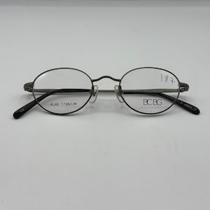 May include: Oval-shaped eyeglasses with a dark gray metal frame. The lenses are clear, and the temples are black. The text "PURE TITANIUM" is printed on the frame. The brand name "BCBG" is also visible.