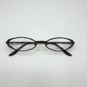 May include: Black oval-shaped eyeglasses with clear lenses. The frames are dark and feature decorative accents on the outer edges. The brand name "Vera Wang" is visible on the arm.