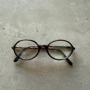 May include: Oval-shaped eyeglasses with a burgundy and black speckled pattern. The frames have a thin metal bridge and temples. The lenses are clear, and the text "DEMO LENSES ONLY" is visible on one lens. The brand name is printed on the temple.
