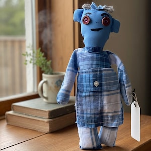 May include: A handmade fabric doll with a light blue head, dark red button eyes, and stitched facial features. The doll wears a blue and white plaid shirt and matching overalls with a single white button on the front pocket and a blank white tag.