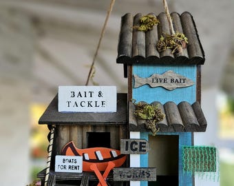 May include: A wooden birdhouse shaped like a bait and tackle shop with a blue and brown exterior. It features signs reading BAIT & TACKLE, LIVE BAIT, BOATS FOR RENT, ICE, WORMS, and OPEN, decorated with a miniature orange canoe, oars, and green moss.