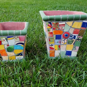 Può includere: Due vasi quadrati a mosaico, uno più grande dell'altro, sono seduti sull'erba verde. I vasi sono fatti di piastrelle di ceramica colorate e hanno un bordo verde.