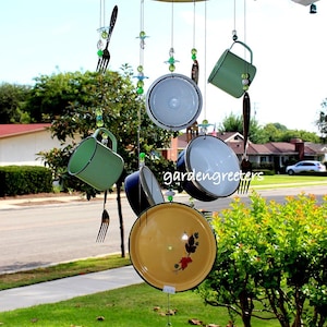 May include: A vintage-style wind chime made from repurposed kitchenware. It features a large yellow enamel lid, smaller enamel lids, mugs, and forks hanging from a metal frame. Green and clear glass beads are strung throughout the chime.