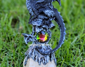 May include: A black dragon statue with iridescent wings perched atop a skull with a jewel in its forehead. The statue is on a bed of green grass.