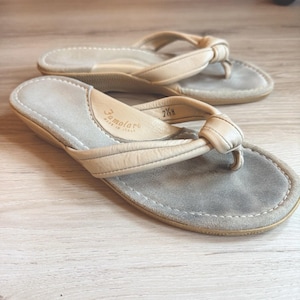 May include: A pair of beige leather thong sandals with a knotted strap design and a grey suede footbed. The interior heel area features the embossed text Famolare Made in Italy and the size 7 1/2 M. The sandals have a low wedge heel profile.