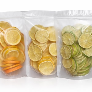 May include: Three clear stand-up pouches containing dehydrated fruit slices. The left pouch holds orange slices, the middle pouch contains yellow lemon slices, and the right pouch is filled with green lime slices, all visible through the plastic.