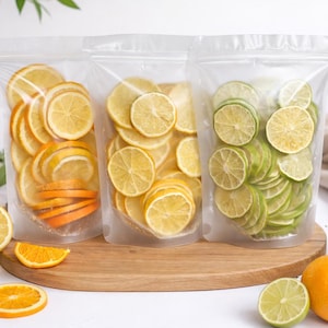 May include: Three clear bags filled with dried citrus slices: oranges, lemons, and limes. The bags sit on a wooden board, with fresh citrus fruits and flowers scattered around. A visually appealing display of ingredients.