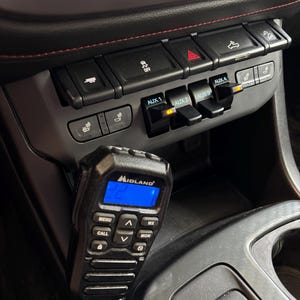 May include: A black Midland radio with a blue display is mounted in a vehicle's console. The console features black buttons and switches, including AUX 1, AUX 2, AUX 3, and AUX 4. The interior has black and gray elements with red stitching.