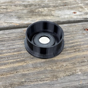 May include: A black, 3D-printed object with a circular design. The object has a wide, ridged outer ring and a smaller, recessed inner circle with a silver disc in the center. The item is sitting on a weathered wooden surface.