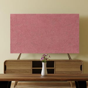 Op de afbeelding: Een groot, rechthoekig, stoffig roze paneel met een herhalend reliëf madeliefpatroon. Het paneel wordt ondersteund door twee schuine, zilverkleurige poten. Onder het paneel staat een houten kast met een vaas bloemen.