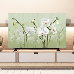 Puede incluir: Una pantalla de televisión muestra una pintura de orquídeas blancas con tallos y hojas verdes sobre un fondo verde suave. Una pequeña mariposa amarilla está posada en un tallo. El televisor está sobre un mueble de madera clara con cajones blancos.