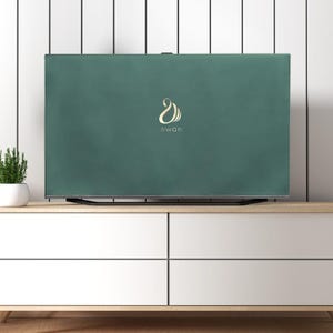 以下が含まれることがあります： ゴールドの筆記体で「swan」と書かれた濃い緑色のテレビカバー。カバーは長方形で、テレビ画面全体を覆っています。カバーは白いキャビネットの上にあり、白い鉢に入った小さな植物があります。