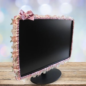 Peut inclure: Un cache-écran décoratif avec une bordure en satin rose et des détails en dentelle. Le cache-écran est orné d'un nœud rose avec un accent de perle sur le dessus et des ornements floraux sur les côtés. Le moniteur est sur un support noir.