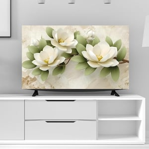 Op de afbeelding: Een flatscreen-tv toont een bloemenafbeelding van witte magnoliabloesems met gele centra en groene bladeren. De bloemen zijn gerangschikt op een bruine tak tegen een getextureerde, neutrale achtergrond. De tv staat op een witte kast.