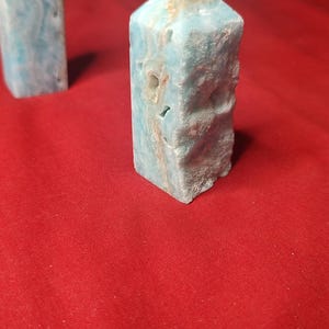 May include: Two light blue gemstone obelisks with natural brown veining. The obelisks are rectangular with a pointed top. They are set against a red background.