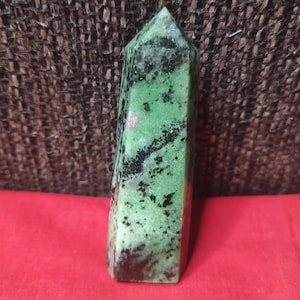 Peut inclure: Un obélisque en pierre précieuse verte et noire avec un sommet pointu. La pierre a un aspect moucheté avec des zones roses et blanches. L'obélisque est posé sur une surface rouge.