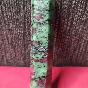 May include: A tall, hexagonal crystal point. The stone is a mix of green, black, and red hues. The crystal is standing upright against a textured brown background and a red surface.