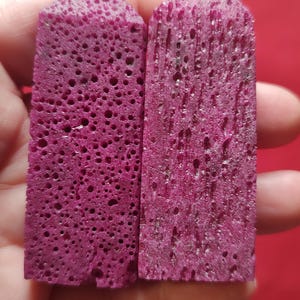 May include: Two magenta-colored, rectangular crystal towers with a porous texture. The crystals are held in a hand against a red background. The crystals have a rough, uneven surface and are cut into a pointed shape at the top.