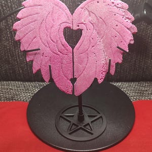May include: A pink, heart-shaped sculpture of angel wings, mounted on a black metal stand. The wings have detailed feather-like textures and a heart-shaped cutout in the center. The base is a black circle with a star design.