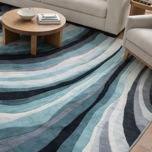 May include: A large area rug featuring a wavy, multi-tonal pattern in shades of blue, teal, gray, and white. The design consists of flowing, curved bands of varying widths that create a sense of movement across the rectangular floor covering.