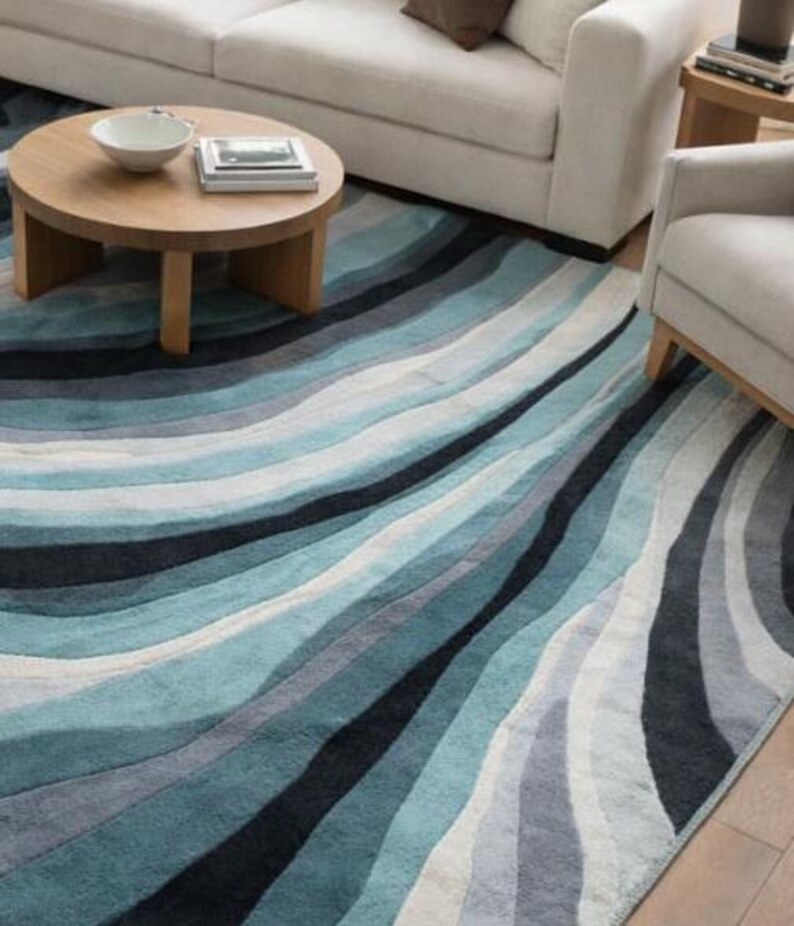 May include: A large area rug featuring a wavy, multi-tonal pattern in shades of blue, teal, gray, and white. The design consists of flowing, curved bands of varying widths that create a sense of movement across the rectangular floor covering.
