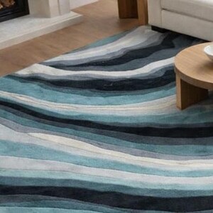 May include: A rectangular area rug featuring a layered, wavy pattern in shades of navy blue, teal, light blue, gray, and white. The design consists of horizontal, undulating bands that create a fluid, multi-toned visual effect across the surface.