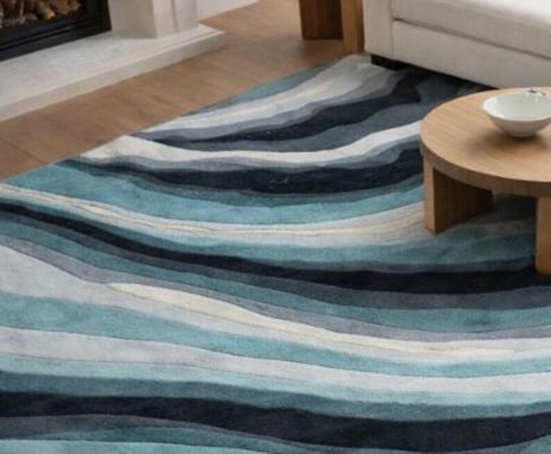 May include: A rectangular area rug featuring a layered, wavy pattern in shades of navy blue, teal, light blue, gray, and white. The design consists of horizontal, undulating bands that create a fluid, multi-toned visual effect across the surface.