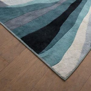 May include: A rectangular area rug featuring a series of flowing, wave-like bands in shades of deep black, dark teal, muted blue, light gray, and off-white. The rug has a finished edge binding and a soft, textured pile surface with a repeating pattern.