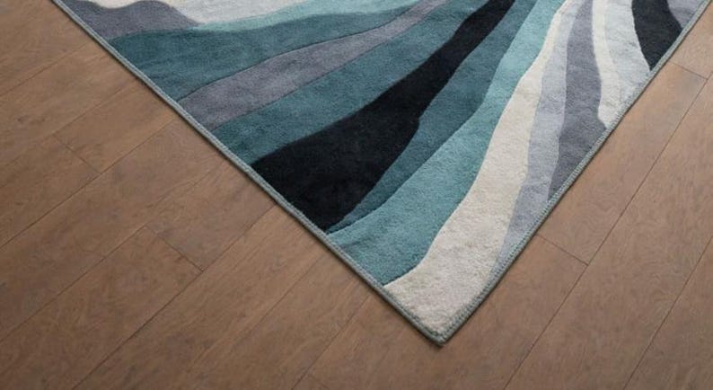 May include: A rectangular area rug featuring a series of flowing, wave-like bands in shades of deep black, dark teal, muted blue, light gray, and off-white. The rug has a finished edge binding and a soft, textured pile surface with a repeating pattern.