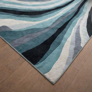 May include: A rectangular area rug featuring a flowing, wavy pattern of layered bands in shades of deep black, dark teal, light blue, grey, and off-white. The rug has a soft, textured pile surface with clean, finished edges along the perimeter.