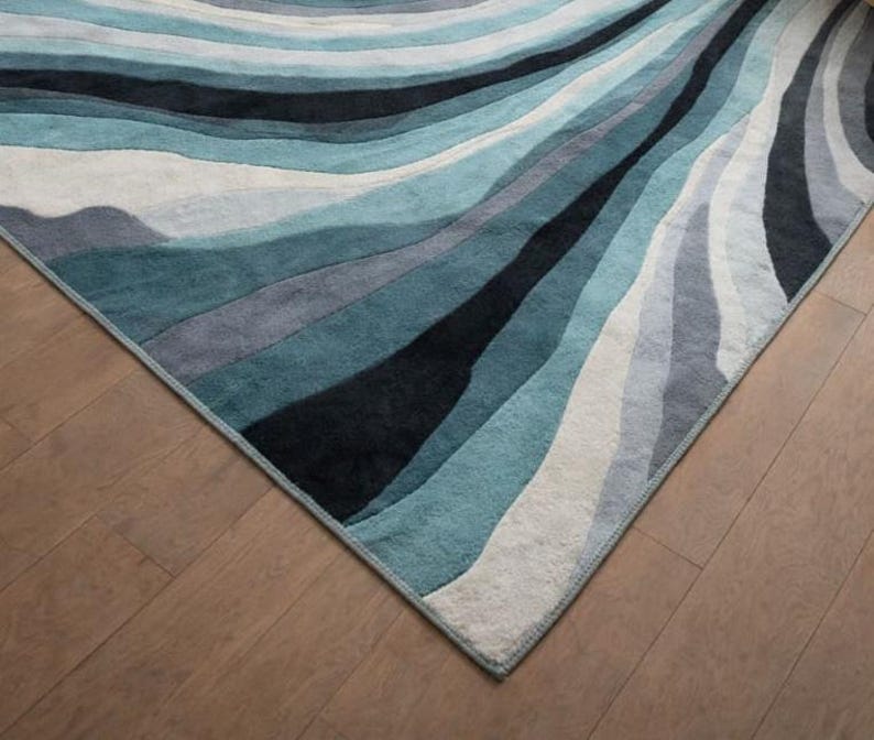 May include: A rectangular area rug featuring a flowing, wavy pattern of layered bands in shades of deep black, dark teal, light blue, grey, and off-white. The rug has a soft, textured pile surface with clean, finished edges along the perimeter.