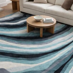 May include: A large rectangular area rug featuring a flowing, wavy pattern in various shades of blue, teal, navy, gray, and white. The design consists of undulating horizontal bands that create a sense of movement across the entire surface.